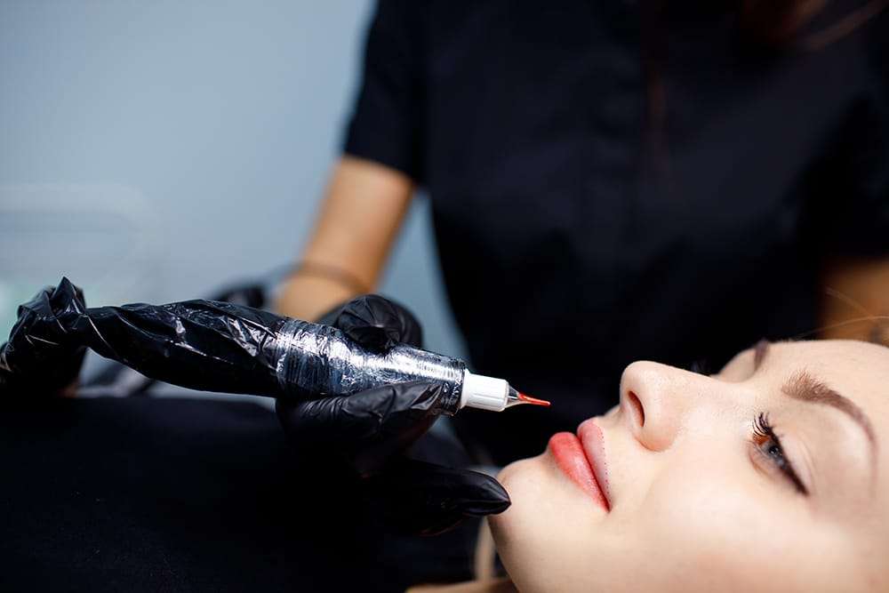 Cosmetologist applying red permanent make up tattoo on young woman lips. Permanent lips tattoo procedure concept.